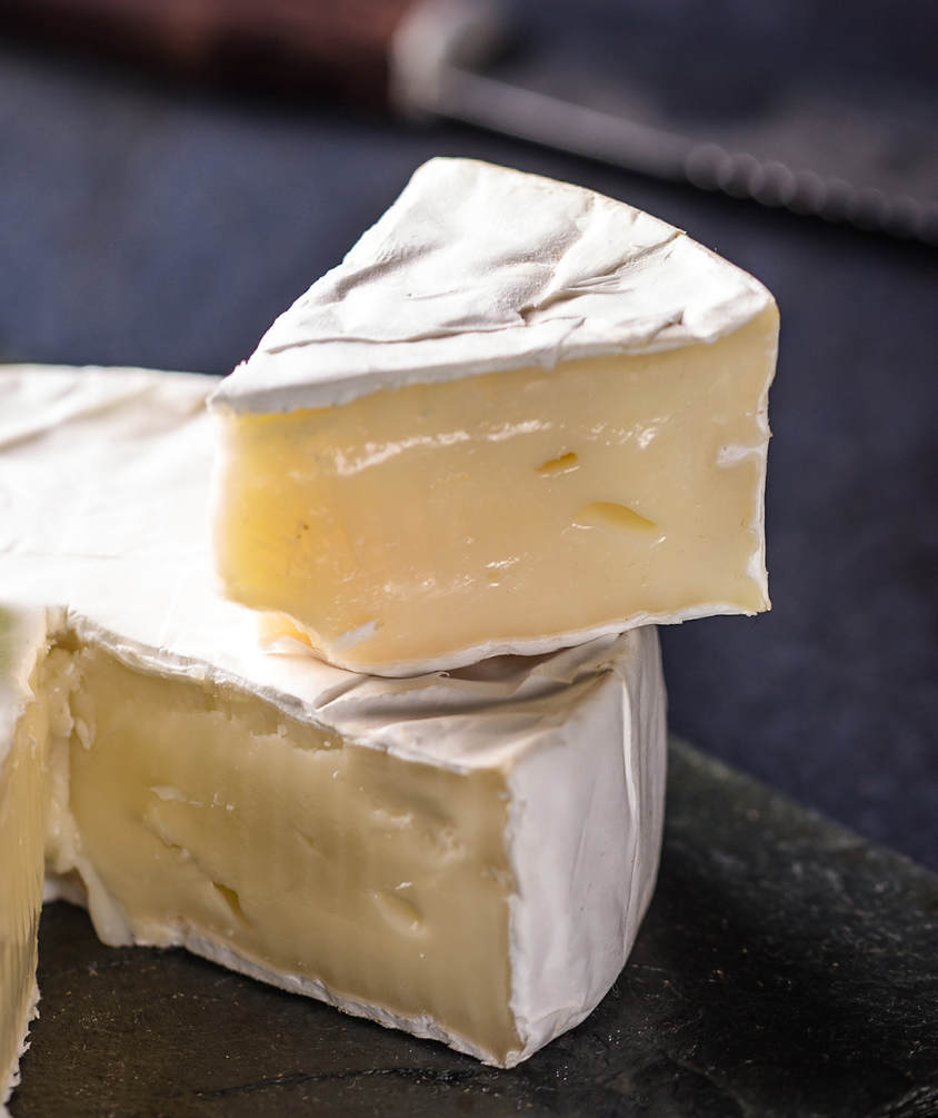 A round wheel of MouCo Camembert with a bloomy white rind, with a wedge cut to reveal its soft, creamy cheese inside.