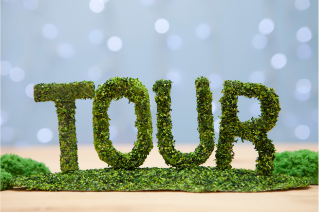 The word “TOUR” formed from textured green moss or foliage, standing upright on a wooden surface with a soft, blurred light background.
