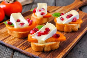 Crostini with persimmons, pomegranates, and MouCo Camembert on a wood platter.